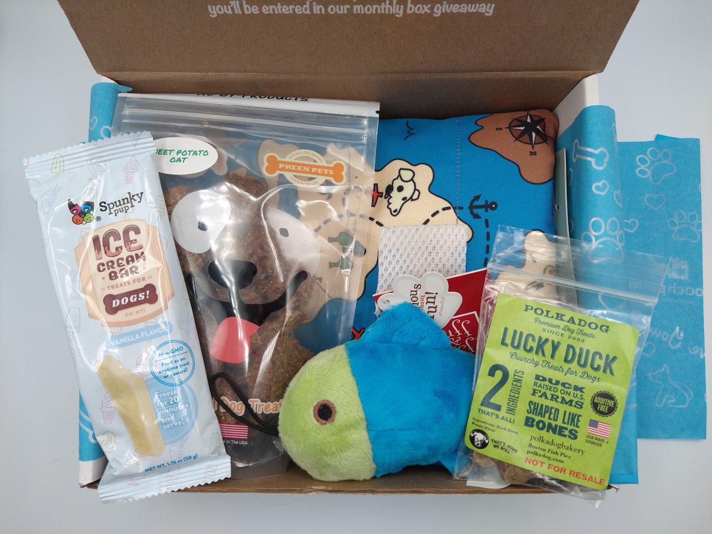 Sample Pooch Perks Box with toys in the shape of a fish and map. The treats are duck, oat and an ice cream treat for the freezer